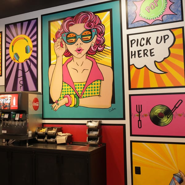 Pop art murals with a woman in sunglasses, headphones, and "Pick Up Here" sign above a soda fountain.